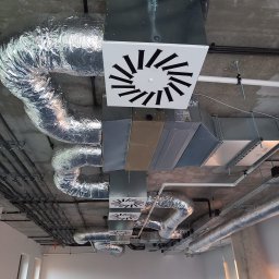 SYSCON - Instalacja wentylacyjna z rekuperacją pod betonowym sufitem, widoczne rury aluminiowe i skrzynki wentylacyjne z czarnymi wlotami powietrza.