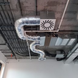 SYSCON - Widok instalacji wentylacyjnych pod sufitem w surowym wnętrzu, widoczne srebrne rury izolacyjne, czarne kable, kratki wentylacyjne i betonowy sufit.