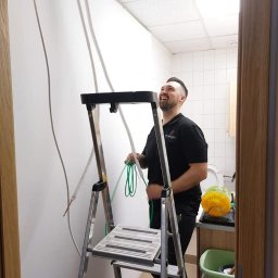 SYSCON - Instalator na drabinie, uśmiechnięty, montuje przewody w pomieszczeniu z kasetonowym sufitem i widoczną toaletą w tle.