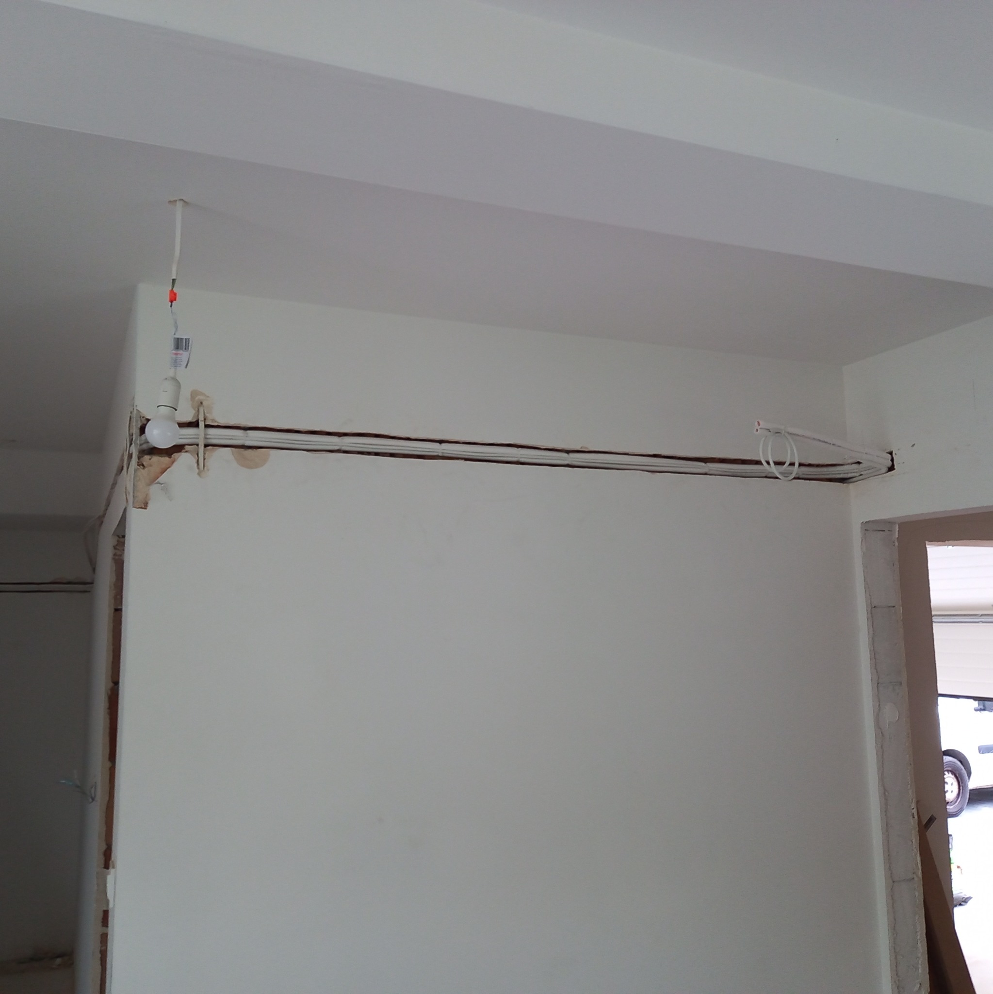 Biała ściana w trakcie bruzdowania pod instalację, widoczne kable i prowizoryczne oświetlenie z żarówki, etap prac w domu jednorodzinnym.