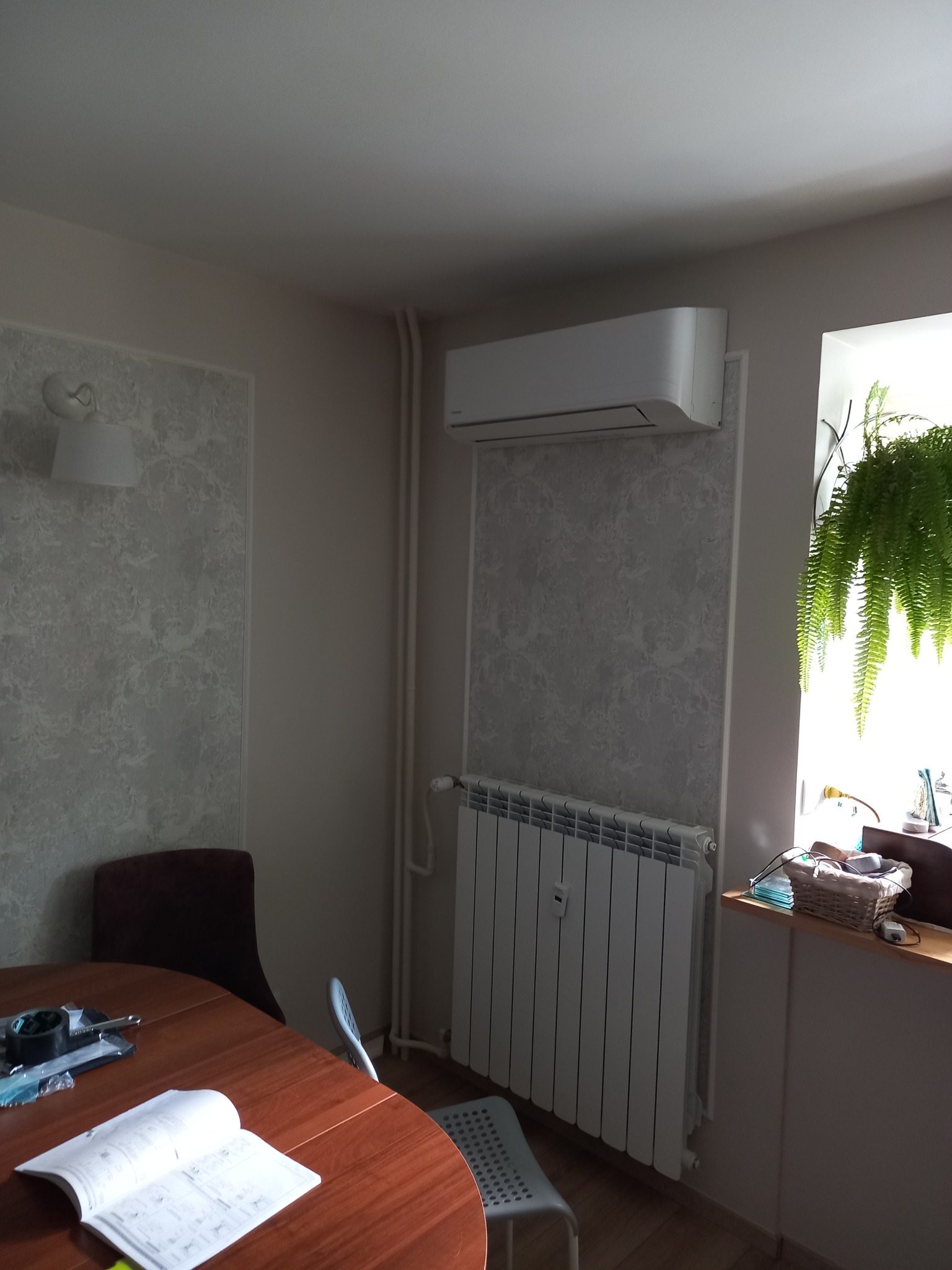 Zamontowana jednostka klimatyzacji ściennej nad grzejnikiem w pokoju z wzorzystą tapetą i wiszącą rośliną w oknie, na stole leży otwarta instrukcja obsługi.