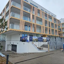 Nowoczesny budynek apartamentowy z drewnianymi elementami elewacji i szklanymi balkonami, widok z poziomu ulicy, z kontenerem budowlanym i zaparkowanym samochodem w pobliżu.