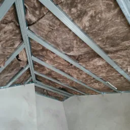 Skośny sufit poddasza w trakcie prac: wełna mineralna między metalowymi profilami, ściany przygotowane do dalszych prac wykończeniowych.