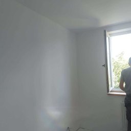 Tom-Bud Sebastian Kaczmarek - Świeżo pomalowane na biało wnętrze pokoju z otwartym oknem, widoczne ślady zabezpieczeń na podłodze, fragment sylwetki osoby stojącej przy oknie.