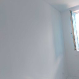 Tom-Bud Sebastian Kaczmarek - Świeżo pomalowane na biało wnętrze pokoju, okno zabezpieczone folią malarską, widoczne wystające przewody elektryczne.