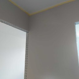 Tom-Bud Sebastian Kaczmarek - Świeżo pomalowana szara ściana z żółtą taśmą malarską przy suficie i metalowym profilem przy drzwiach.