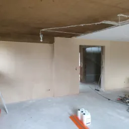 Wnętrze w trakcie prac wykończeniowych: ściany pokryte świeżo nałożoną gładzią, surowy sufit z wystającymi przewodami, na podłodze butelki z gruntem i narzędzia.