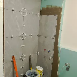 Ściana łazienki w trakcie układania szarych płytek imitujących marmur, z użyciem poziomicy NEO i systemu poziomowania płytek; widoczne przygotowane podłoże i instalacje.