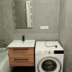 Wykończona łazienka z szarymi płytkami imitującymi marmur, szafką pod umywalką z drewnopodobnymi frontami, czarną armaturą i białą pralką Gorenje.