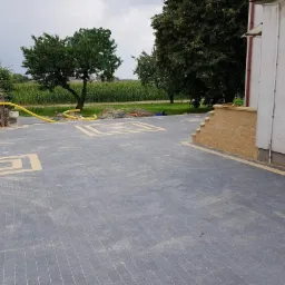 Ułożona kostka brukowa w kolorze szarym z dekoracyjnymi kwadratami w odcieniu piaskowym, żółty wąż leżący na kostce, częściowo ułożone schody z piaskowej kostki przy ścianie budynku.