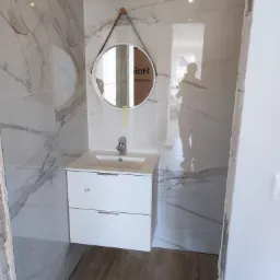 Wykończona łazienka z marmurowymi płytkami, okrągłe lustro na skórzanym pasku nad białą szafką z umywalką, widoczny fragment drewnopodobnej podłogi i odbicie osoby w lustrze.