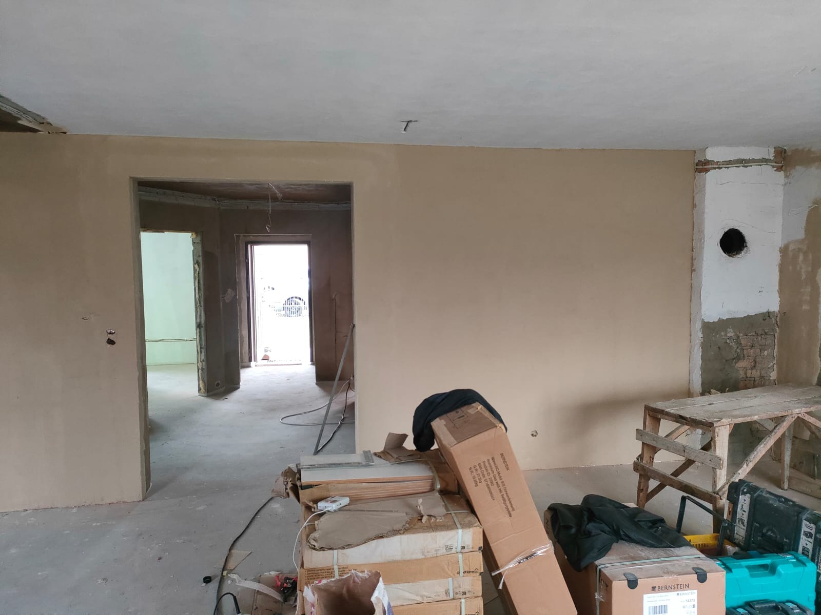 Wnętrze w trakcie remontu: świeżo otynkowane ściany w beżowym odcieniu, widoczne otwory drzwiowe prowadzące do kolejnych pomieszczeń, kartony z materiałami budowlanymi i drewniany stół roboczy...