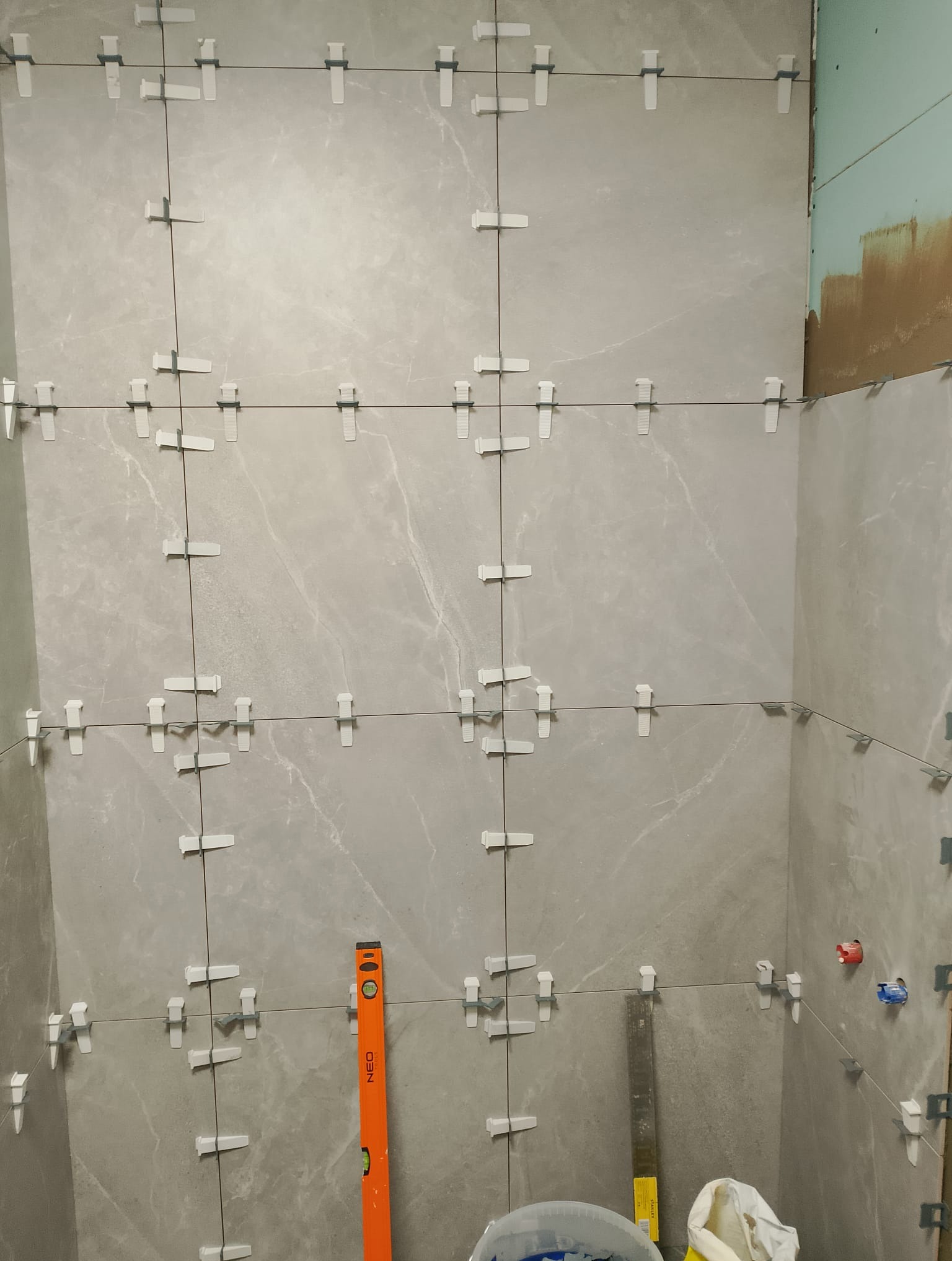 Ściana łazienki w trakcie układania szarych płytek, widoczne krzyżyki dystansowe, sznurek poziomujący i poziomica Neo.