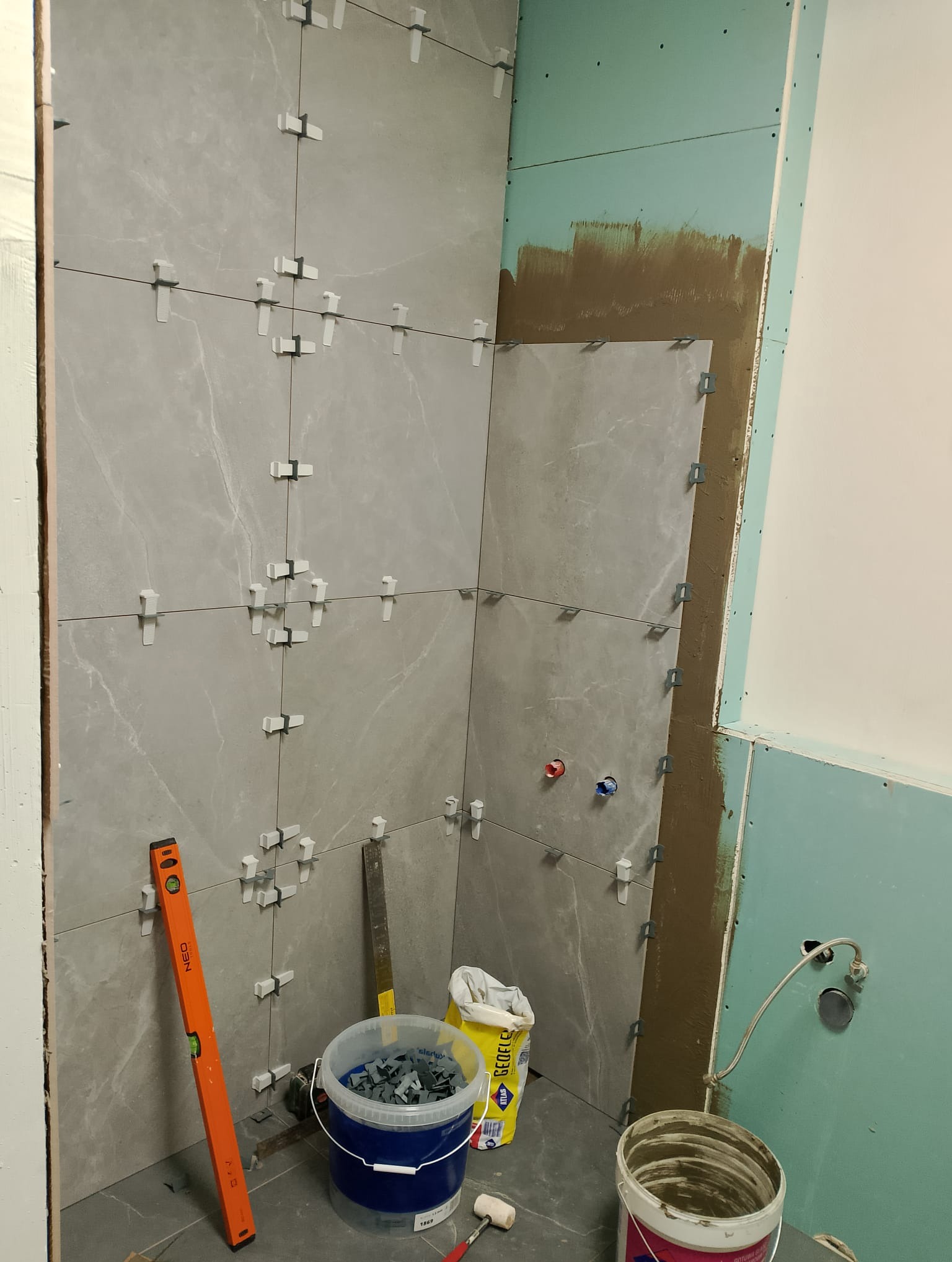 Ściana łazienki w trakcie układania szarych płytek imitujących marmur, z użyciem poziomicy NEO i systemu poziomowania płytek; widoczne przygotowane podłoże i instalacje.