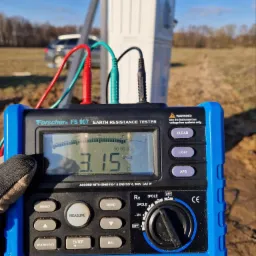 Miernik rezystancji uziemienia Forscher FS 907 z podłączonymi przewodami pomiarowymi, wskazujący wartość 3.15 Ω, na tle szafy elektrycznej i pola.