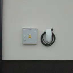 Na elewacji budynku widoczna skrzynka elektryczna z symbolem ostrzegawczym wysokiego napięcia oraz zamontowana stacja ładowania pojazdów elektrycznych marki Tesla z kablem zwiniętym w okrąg.