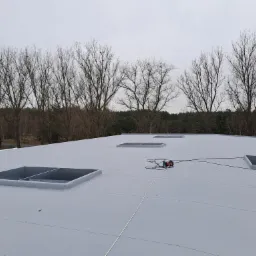 Szary dach płaski z trzema świetlikami, na tle drzew bez liści i pochmurnego nieba. Na dachu leży czarny kabel i czerwono-czarna wiertarka.