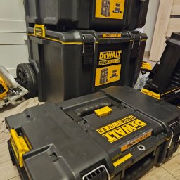 Bartłomiej Cecot Infinity Solution Group - Stos czarnych, modułowych skrzyń narzędziowych DeWalt ToughSystem 2.0 z żółtymi akcentami, ustawionych na podłodze, z widoczną piłą tarczową w tle.