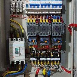 Bartłomiej Cecot Infinity Solution Group - Otwarta skrzynka elektryczna z bezpiecznikami, stycznikami i przewodami o różnych kolorach, zamontowana na ścianie.