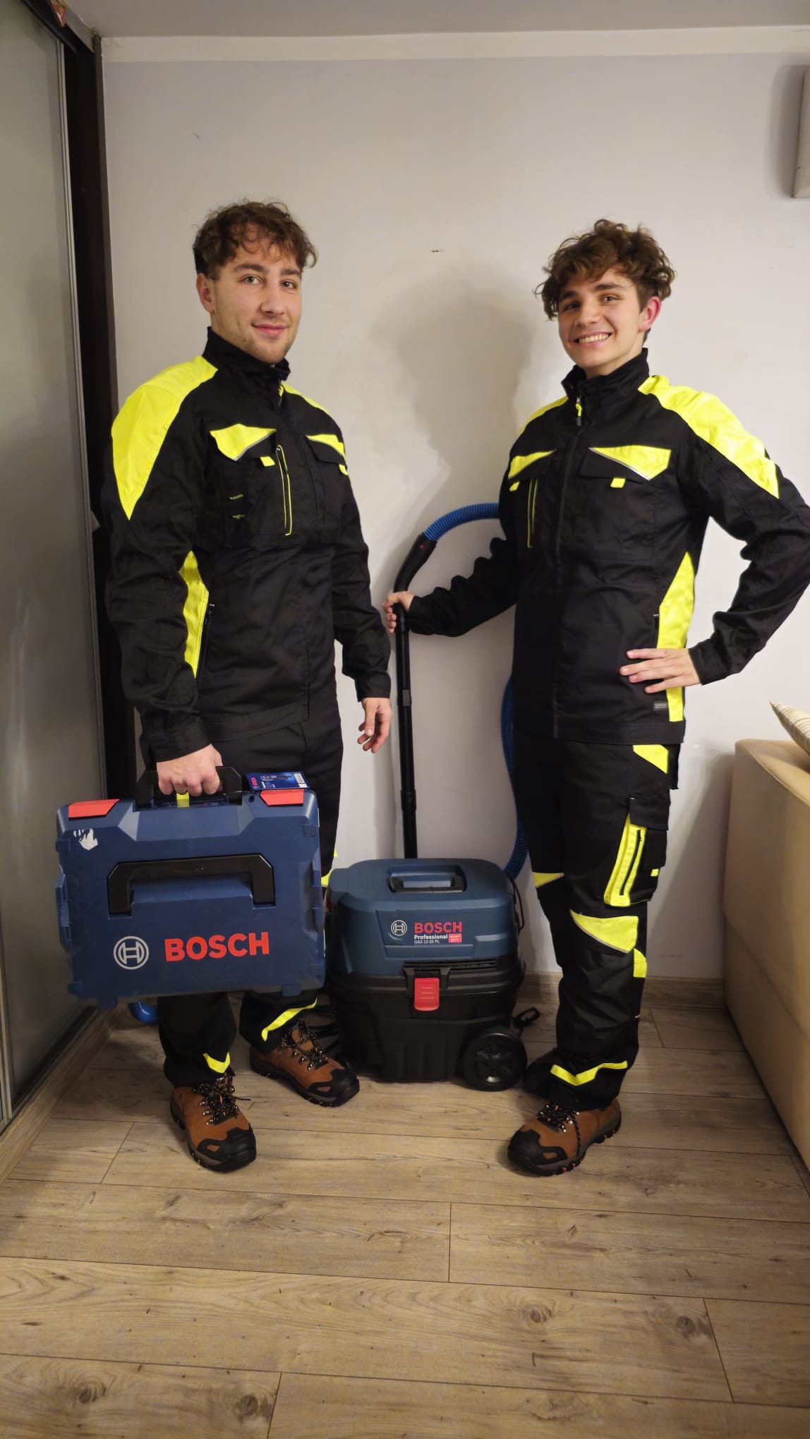 Dwóch młodych mężczyzn w roboczych kombinezonach z żółtymi wstawkami, prezentuje narzędzia marki Bosch: niebieską skrzynkę narzędziową i odkurzacz przemysłowy, stoją na drewnianej podłodze na tle...