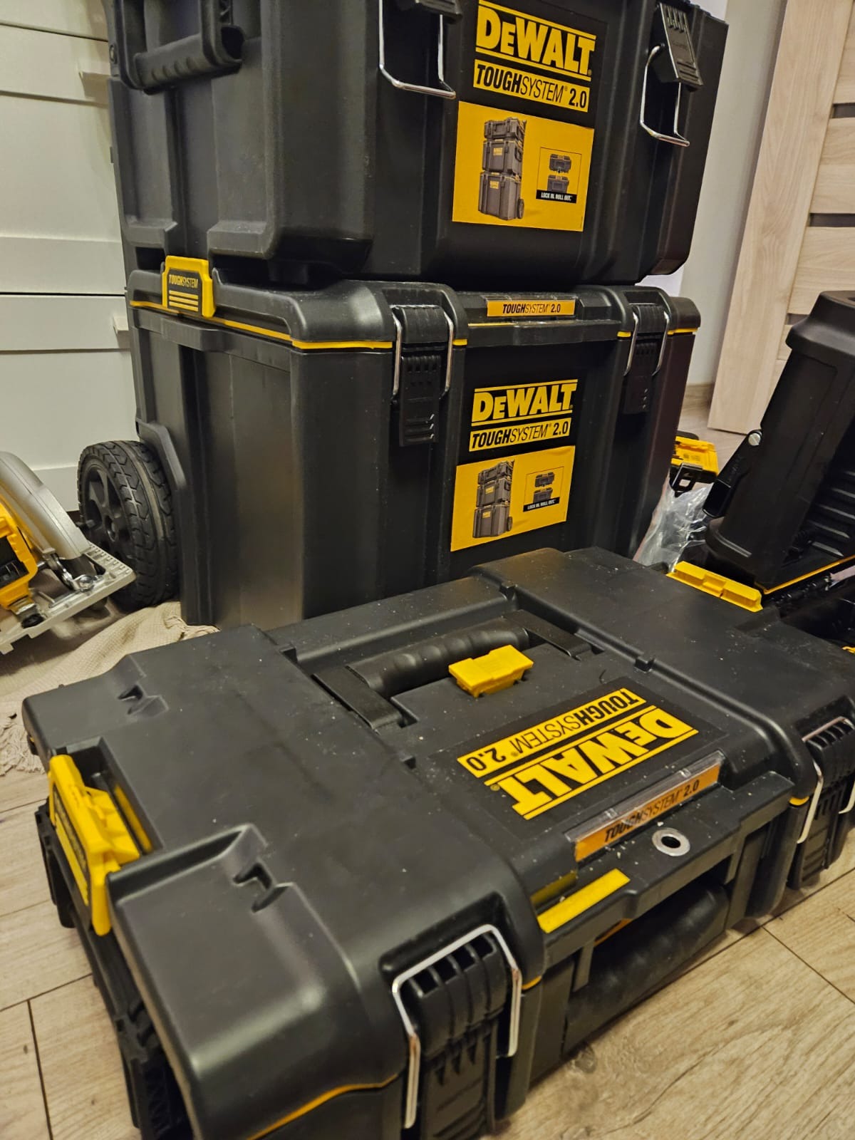 Stos czarnych, modułowych skrzyń narzędziowych DeWalt ToughSystem 2.0 z żółtymi akcentami, ustawionych na podłodze, z widoczną piłą tarczową w tle.