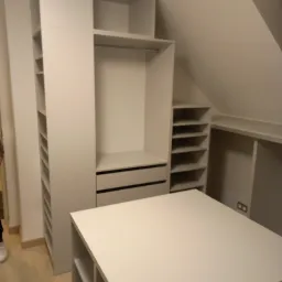 Biała szafa na wymiar z otwartymi półkami i dwiema szufladami, dopasowana do skosu dachu, z dodatkowym modułem z półkami na środku pomieszczenia, widoczne fragmenty podłogi i osoby w tle.