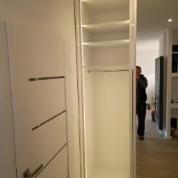 Biała szafa wnękowa z oświetleniem LED, widok na wnętrze z półkami i drążkiem, przesuwne lustrzane drzwi częściowo zasłaniają sylwetkę instalatora.