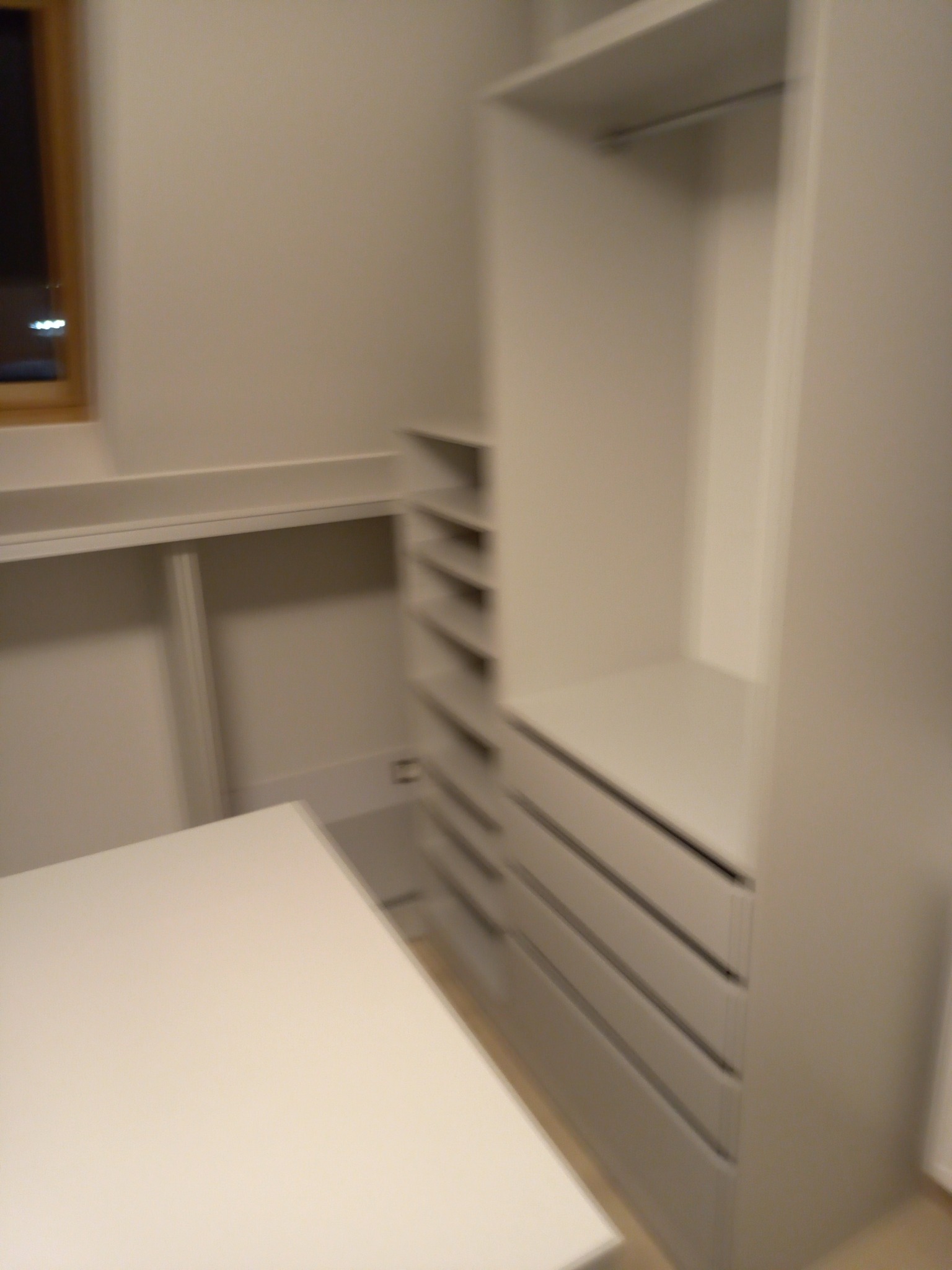 Wnętrze garderoby z białymi meblami: szafa z półkami i szufladami, blat roboczy, widok z boku, delikatne oświetlenie.