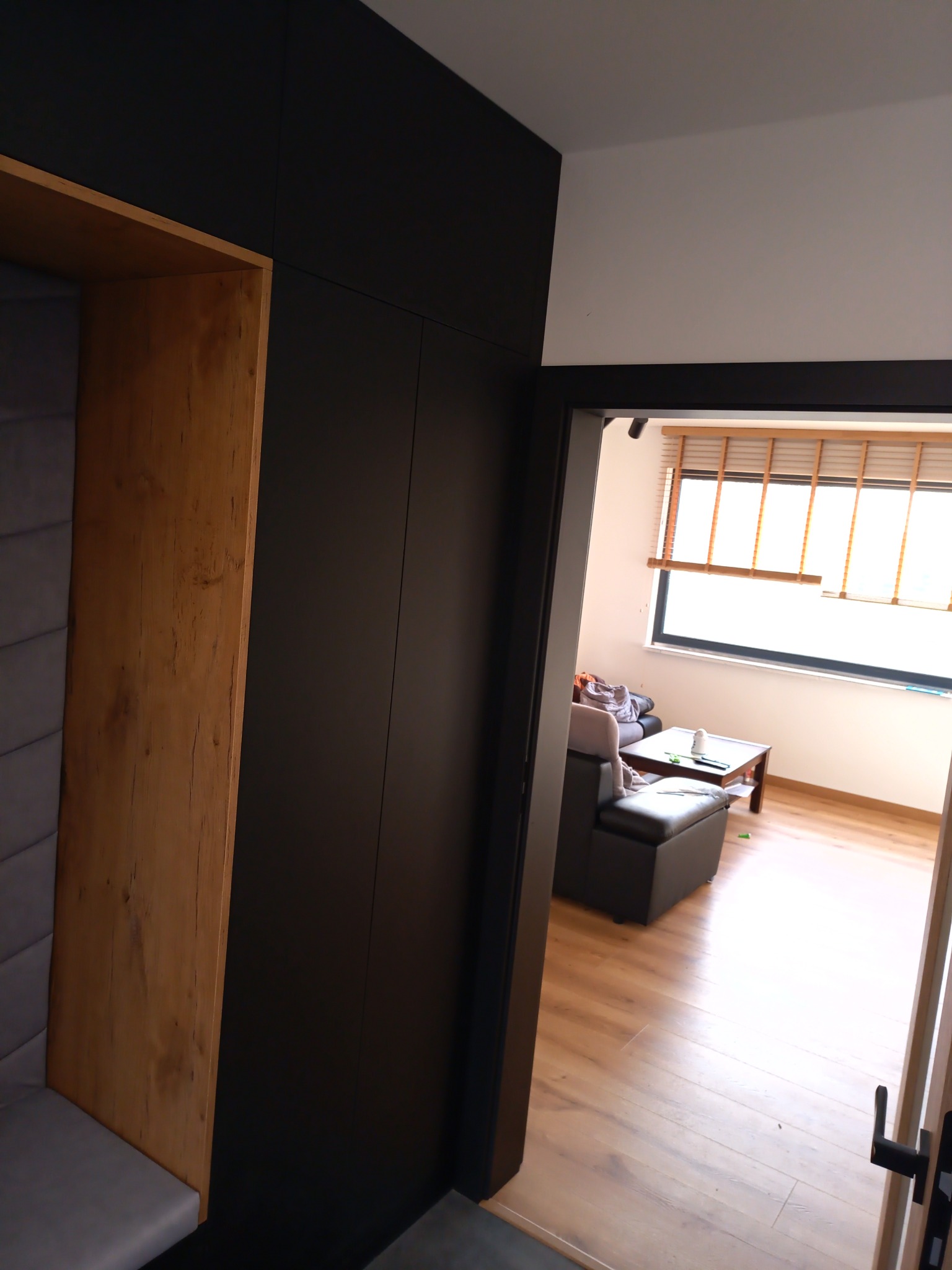 Ciemna szafa na wymiar z wbudowanym drewnianym siedziskiem z szarą tapicerką, widok z perspektywy korytarza na jasny salon z drewnianą podłogą.
