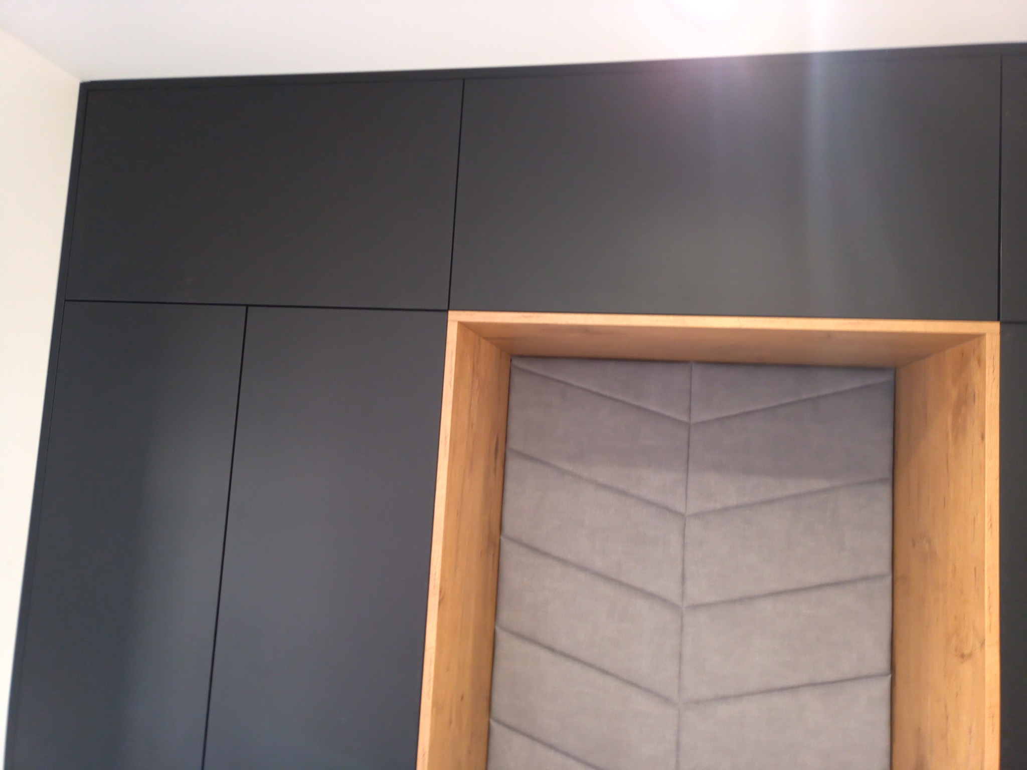 Ciemnoszara zabudowa meblowa z drewnianą wnęką wypełnioną szarą tapicerką z geometrycznym wzorem chevron.