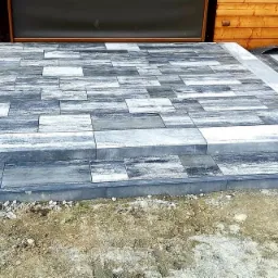 Stopnie wejściowe wyłożone szarą kostką brukową o nieregularnym wzorze, obrzeża z ciemnej kostki, widoczna ubijarka w tle, prace w trakcie realizacji.