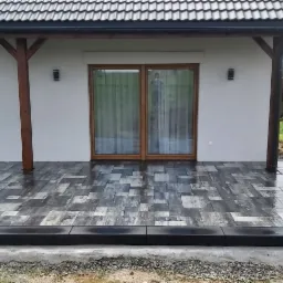 Nowoczesny taras z szarej kostki brukowej o nieregularnym wzorze, zadaszony drewnianą pergolą przy białym domu z przesuwnymi drzwiami balkonowymi, widok na zielony ogród w tle.