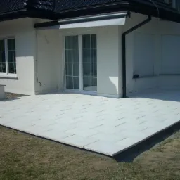 Jasny taras z prostokątnych płyt ułożonych przy białym domu z przesuwnymi drzwiami, widok z poziomu trawnika.