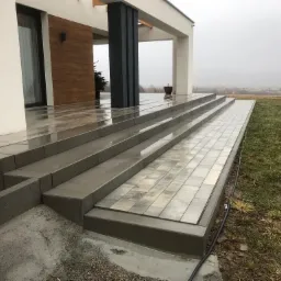 Nowoczesne schody zewnętrzne z szarych płyt prowadzące do domu, widok na trawnik i zamglony krajobraz w tle.