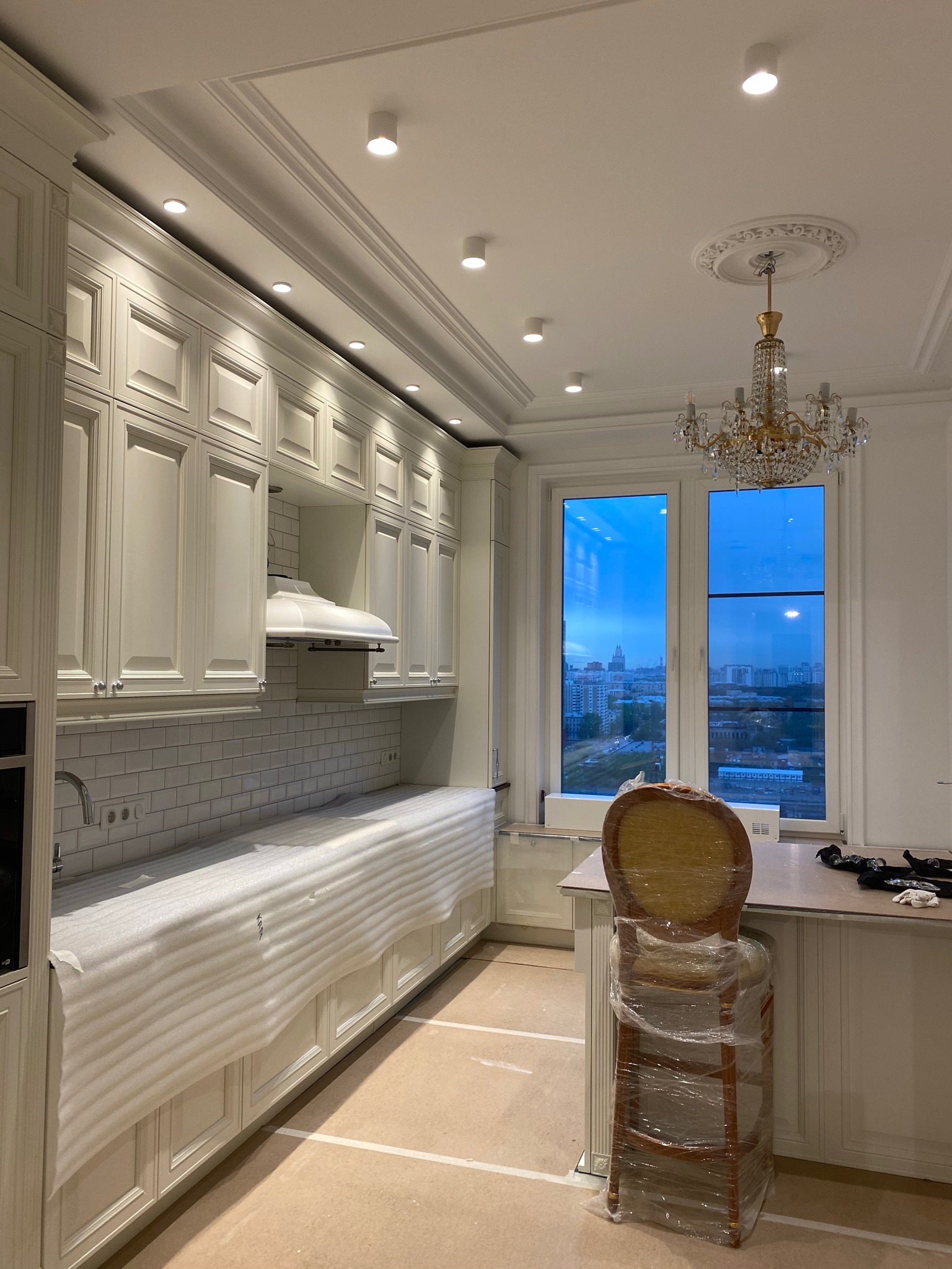 Wnętrze kuchni w trakcie montażu: białe szafki kuchenne z blatem zabezpieczonym folią ochronną, okno z widokiem na miasto, kryształowy żyrandol, krzesło owinięte folią.