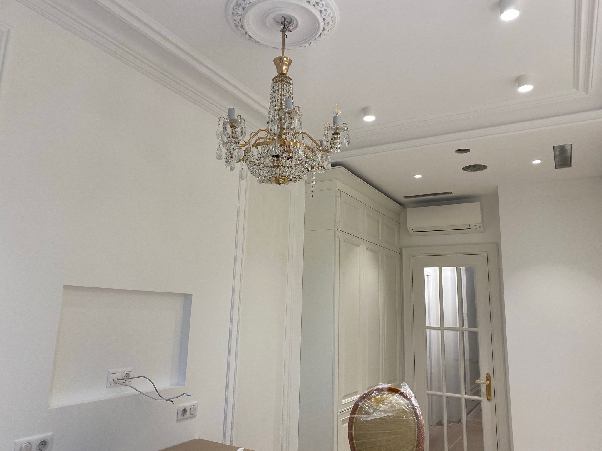 Kryształowy żyrandol zamontowany na suficie w nowym wnętrzu z gładkimi ścianami, widoczne puszki elektryczne i przewody, oraz klimatyzator przy suficie.