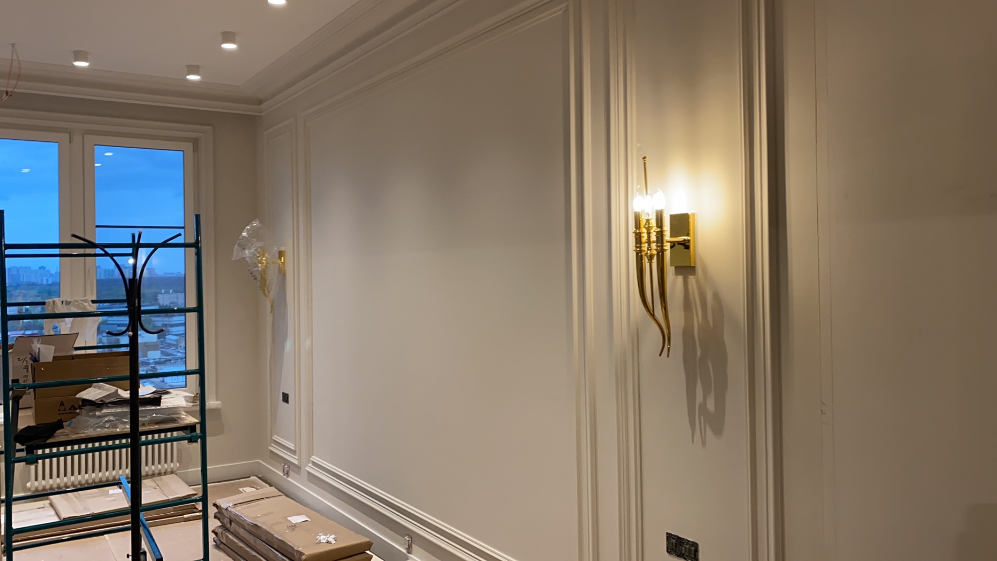 Wnętrze w trakcie wykańczania: dekoracyjne listwy ścienne, złoty kinkiet z trzema żarówkami, widok na miasto z okna, rozstawiony stojak na ubrania i kartony na podłodze.