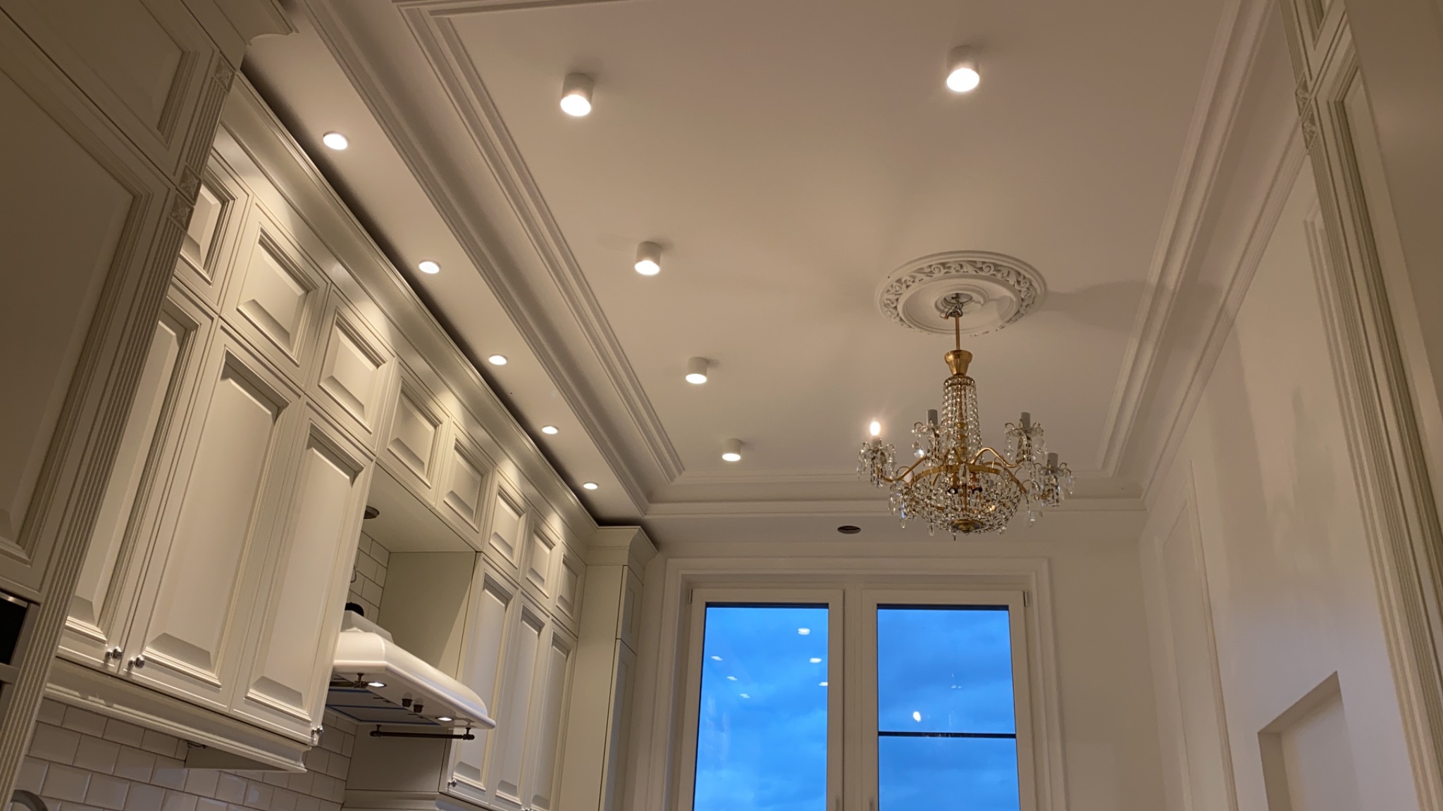 Kryształowy żyrandol zawieszony na suficie z dekoracyjną rozetą, otoczony wpuszczanymi reflektorami i sztukaterią w eleganckiej kuchni z białymi szafkami.