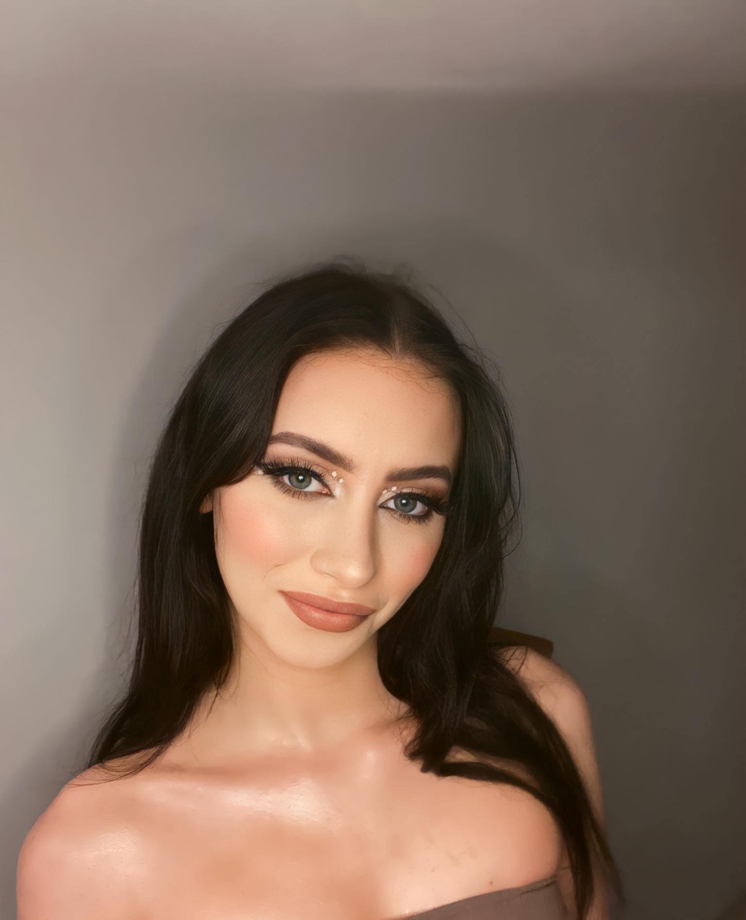 Portret kobiety z ciemnymi włosami i subtelnym, profesjonalnym makijażem z błyszczącymi akcentami wokół oczu, na tle szarej ściany.