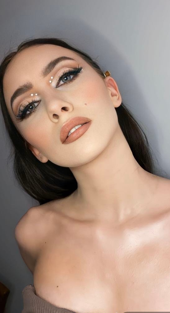 Portret kobiety z delikatnym, beżowym makijażem oczu, sztucznymi rzęsami i ozdobnymi białymi kropkami między brwiami, podkreślone usta w kolorze nude, złota ozdoba we włosach przy uchu.