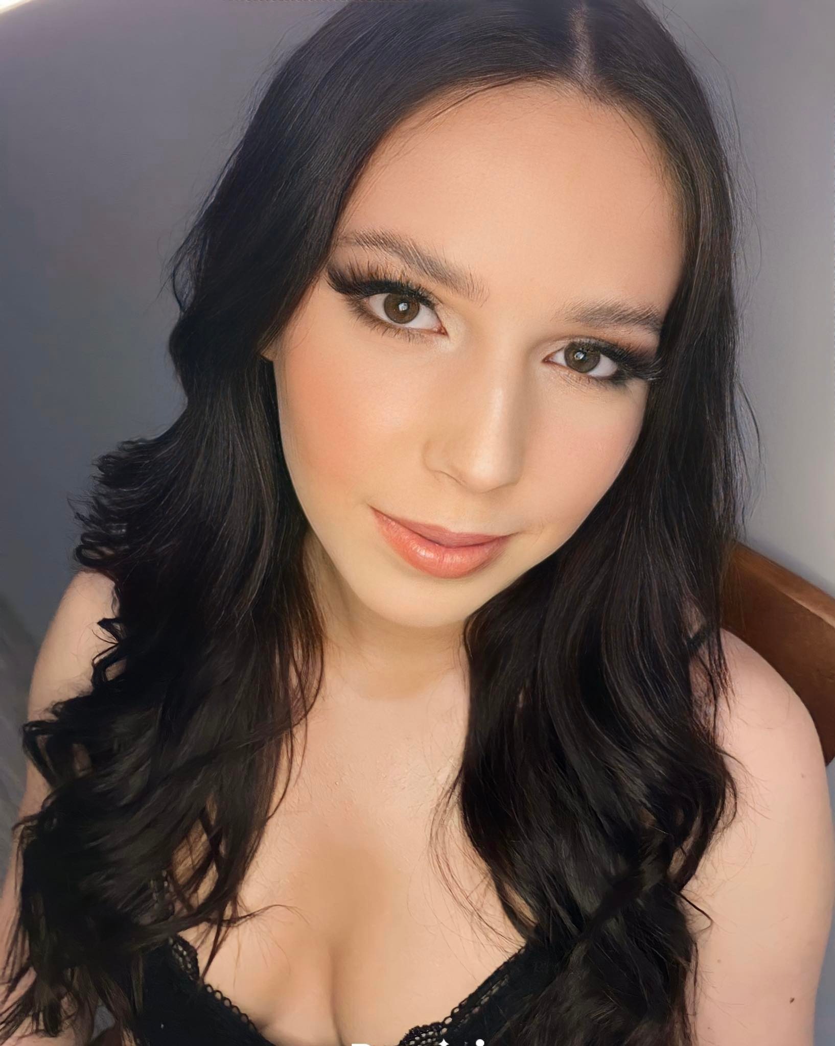 Portret młodej kobiety z delikatnym, naturalnym upiększeniem, podkreślonymi rzęsami i błyszczącymi ustami, z ciemnymi, falującymi włosami.