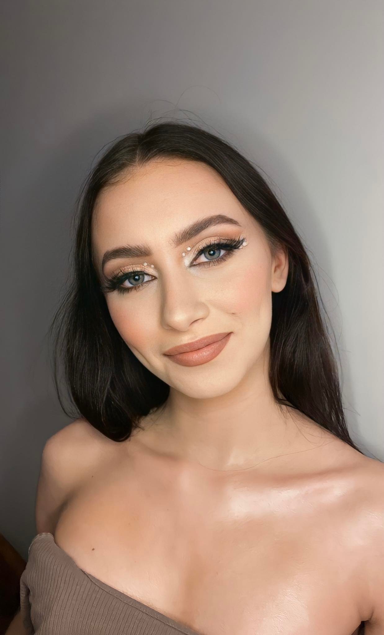 Portret kobiety z delikatnym, beżowym cieniem na powiekach, sztucznymi rzęsami i drobnymi, białymi kryształkami w kącikach oczu, podkreślającymi spojrzenie.