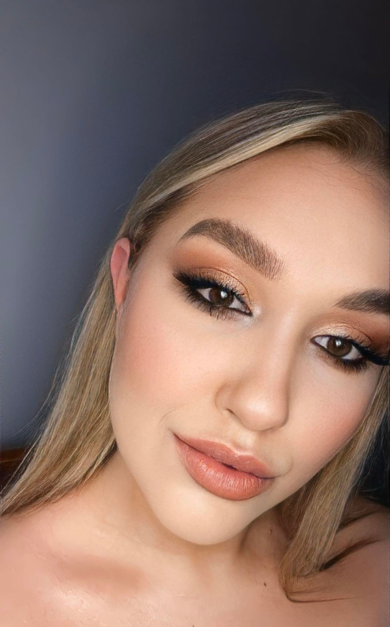 Portret kobiety z profesjonalnie wykonanym, ciepłym, brązowym makijażem oczu i ustami w odcieniu nude, z widocznym przedłużeniem rzęs.