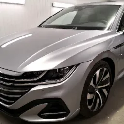 Błyszczący, srebrny Volkswagen Arteon po umyciu, widoczny z przodu pod kątem, z wyraźnymi detalami felg i reflektorów, stojący w jasnym pomieszczeniu z białymi panelami ściennymi.
