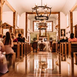 Wnętrze drewnianego kościoła podczas ceremonii ślubnej, widok z tyłu na parę młodą klęczącą przed ołtarzem, goście siedzą w ławkach po obu stronach nawy, podłoga z połyskiem odbija światło...