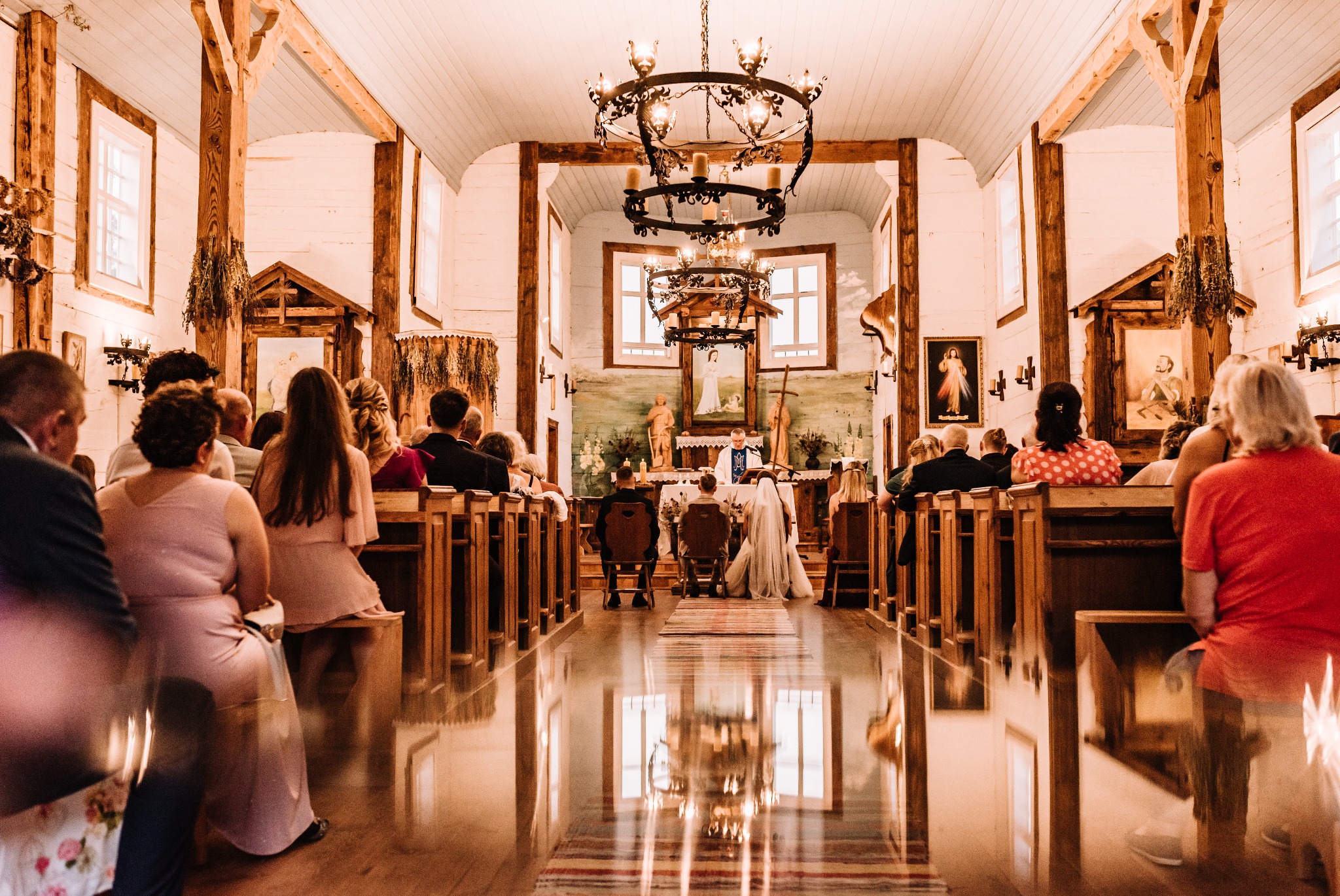 Wnętrze drewnianego kościoła podczas ceremonii ślubnej, widok z tyłu na parę młodą klęczącą przed ołtarzem, goście siedzą w ławkach po obu stronach nawy, podłoga z połyskiem odbija światło...
