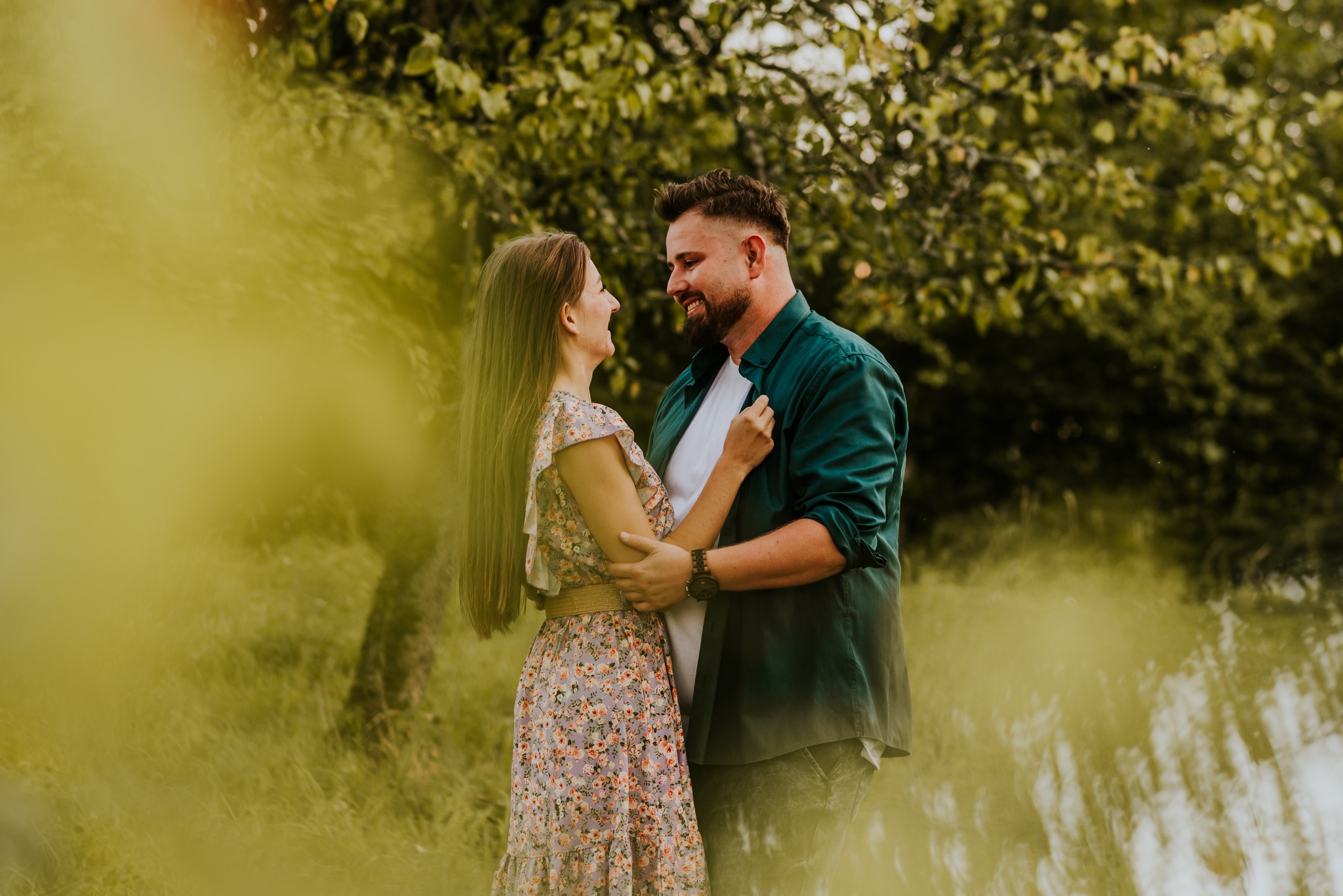 Uśmiechnięta para w objęciach, ona w kwiecistej sukience, on w rozpiętej zielonej koszuli, na tle zieleni i wody.