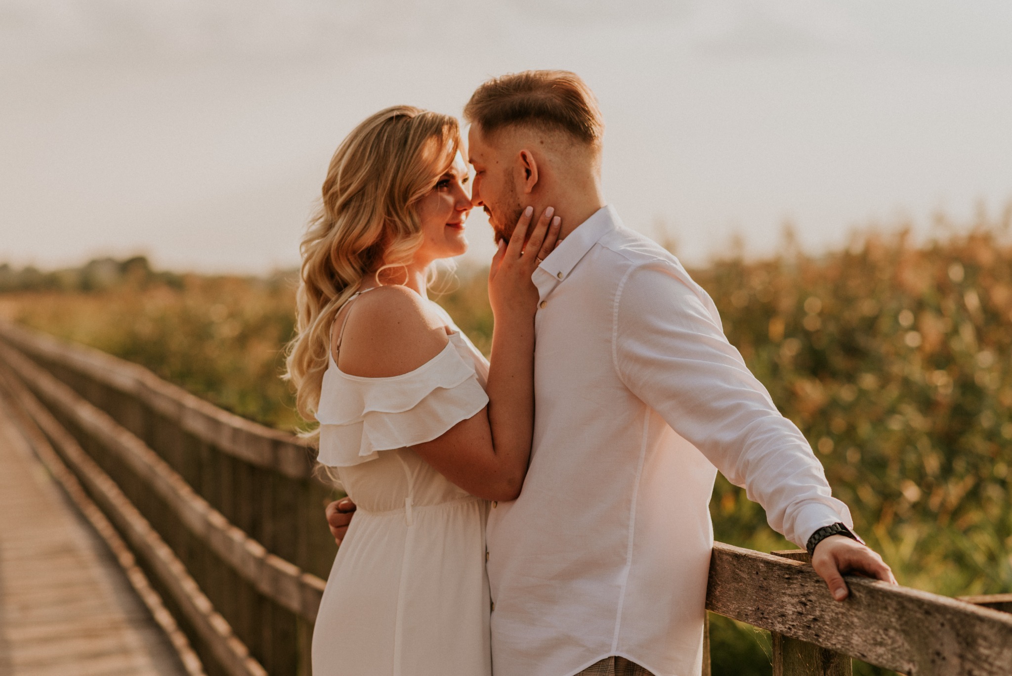 Para zakochanych na drewnianym pomoście w promieniach zachodzącego słońca, kobieta w białej sukience dotyka twarzy mężczyzny.