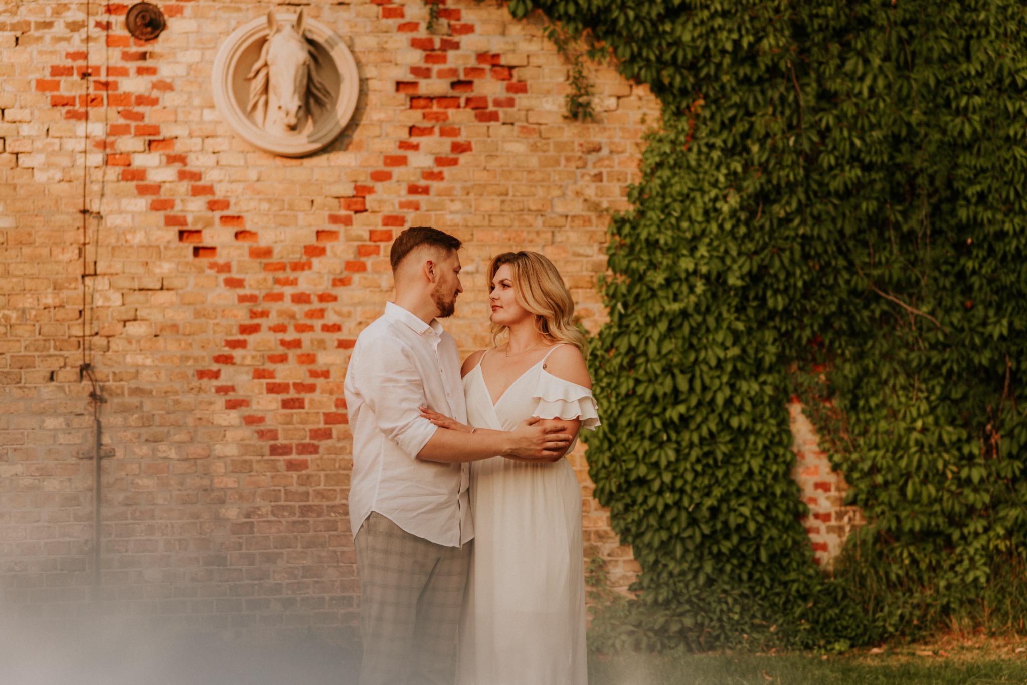 Para narzeczonych obejmuje się na tle ceglanej ściany z rzeźbą końskiej głowy i zielonym bluszczem.