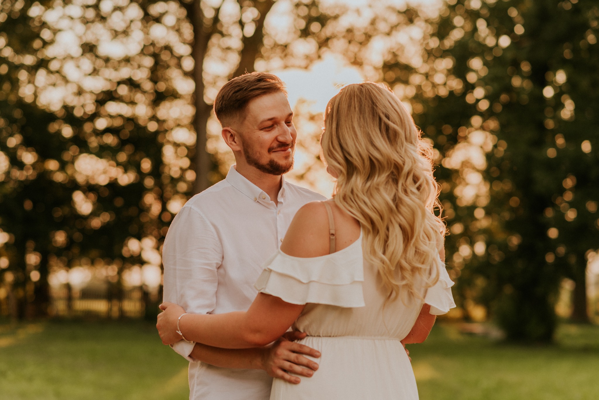 Uśmiechnięta para w objęciach na tle drzew w ciepłym, zachodzącym słońcu; ona w zwiewnej sukience z odkrytymi ramionami, on w białej koszuli z podwiniętymi rękawami.
